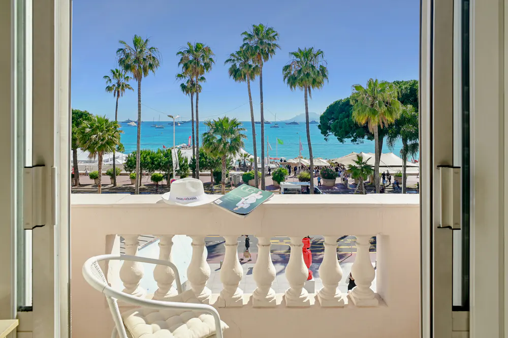 View from a balcony with a white chair, hat, and magazine overlooking a beach with palm trees and turquoise water.