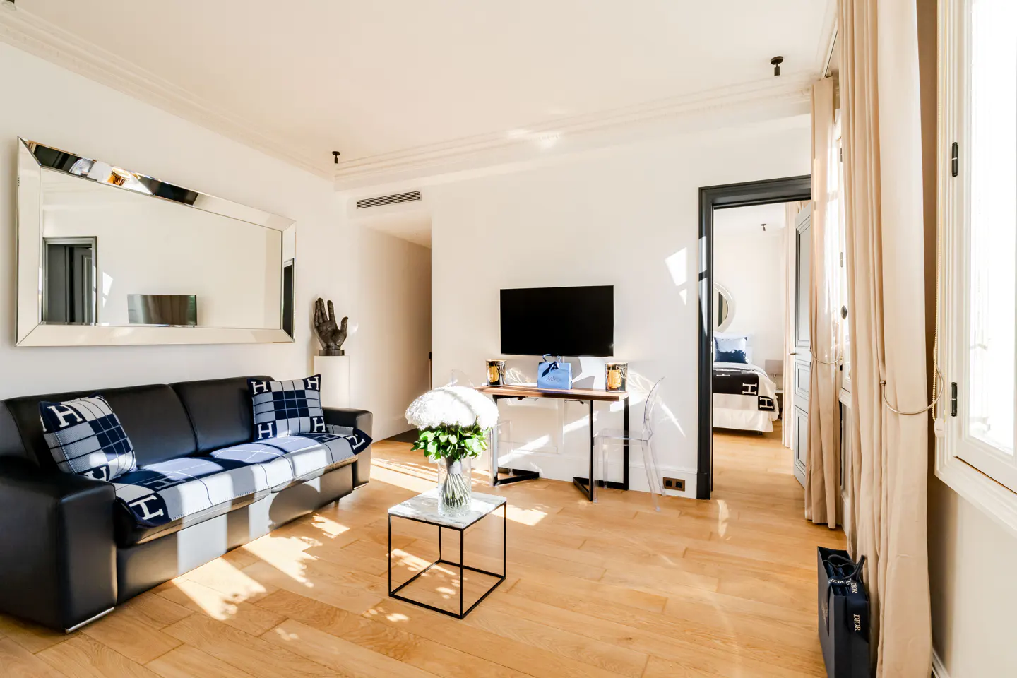 Modern living room with a black leather sofa, white flowers on a marble table, wall-mounted TV, and view into a bedroom.