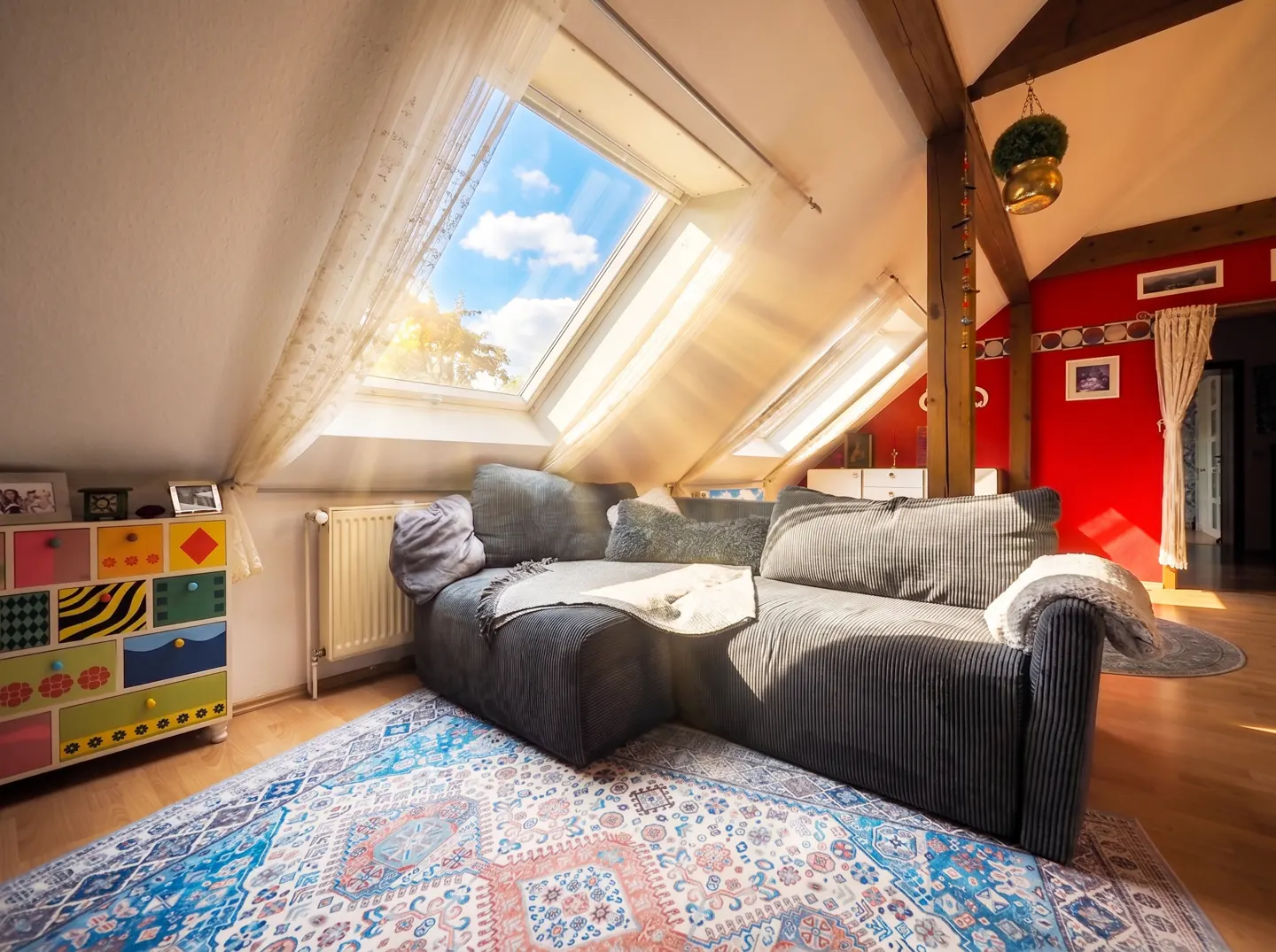 Attic living room with a gray sofa, colorful dresser, and patterned rug. Sunlight streams through skylights with sheer curtains.