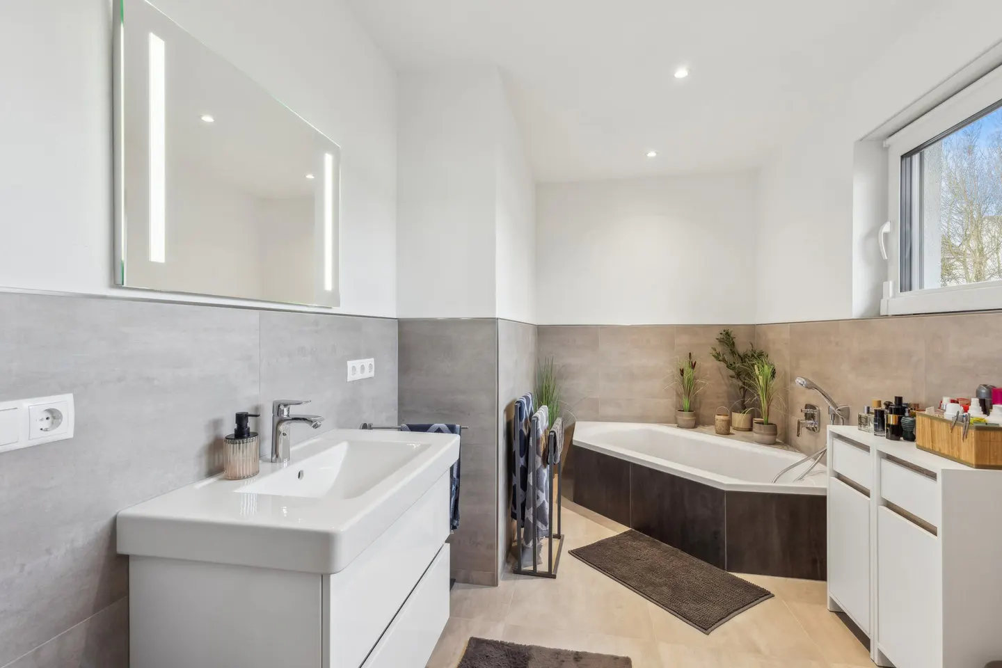 Bright bathroom with white walls, gray tile, and light beige floor. Features a white vanity, tub, and storage cabinet. A window provides natural light.