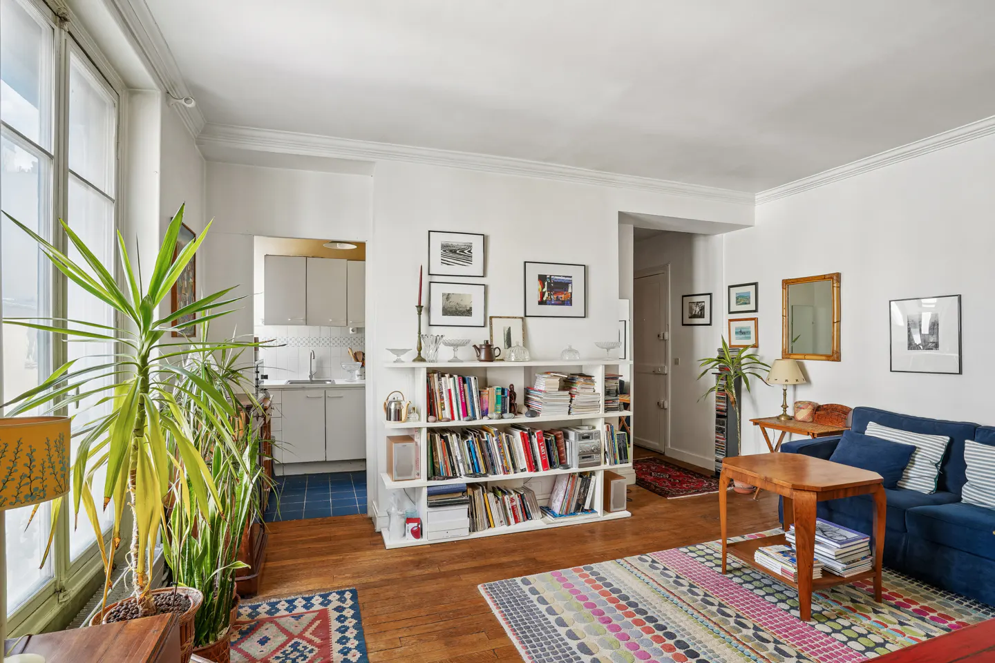 Bright living room with white walls, wood floors, and large windows. A blue sofa, wood table, and colorful rug add warmth. Bookshelves divide the space.