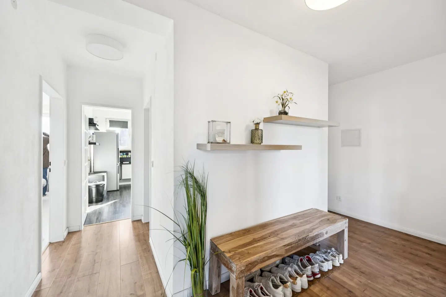 Bright entryway with wood floors, white walls, and a wooden bench with shoes underneath. Shelves hold decor. Hallway leads to kitchen.