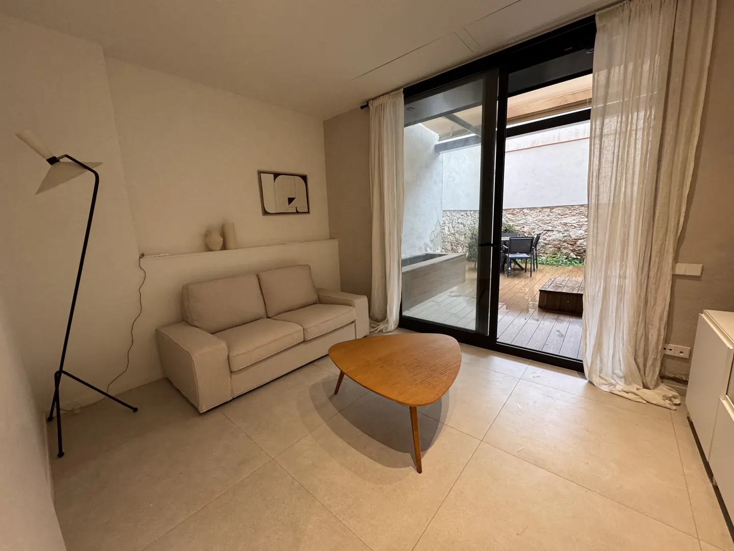 Living room with a beige sofa, wood coffee table, and black floor lamp. Sliding glass doors lead to a patio with stone walls.