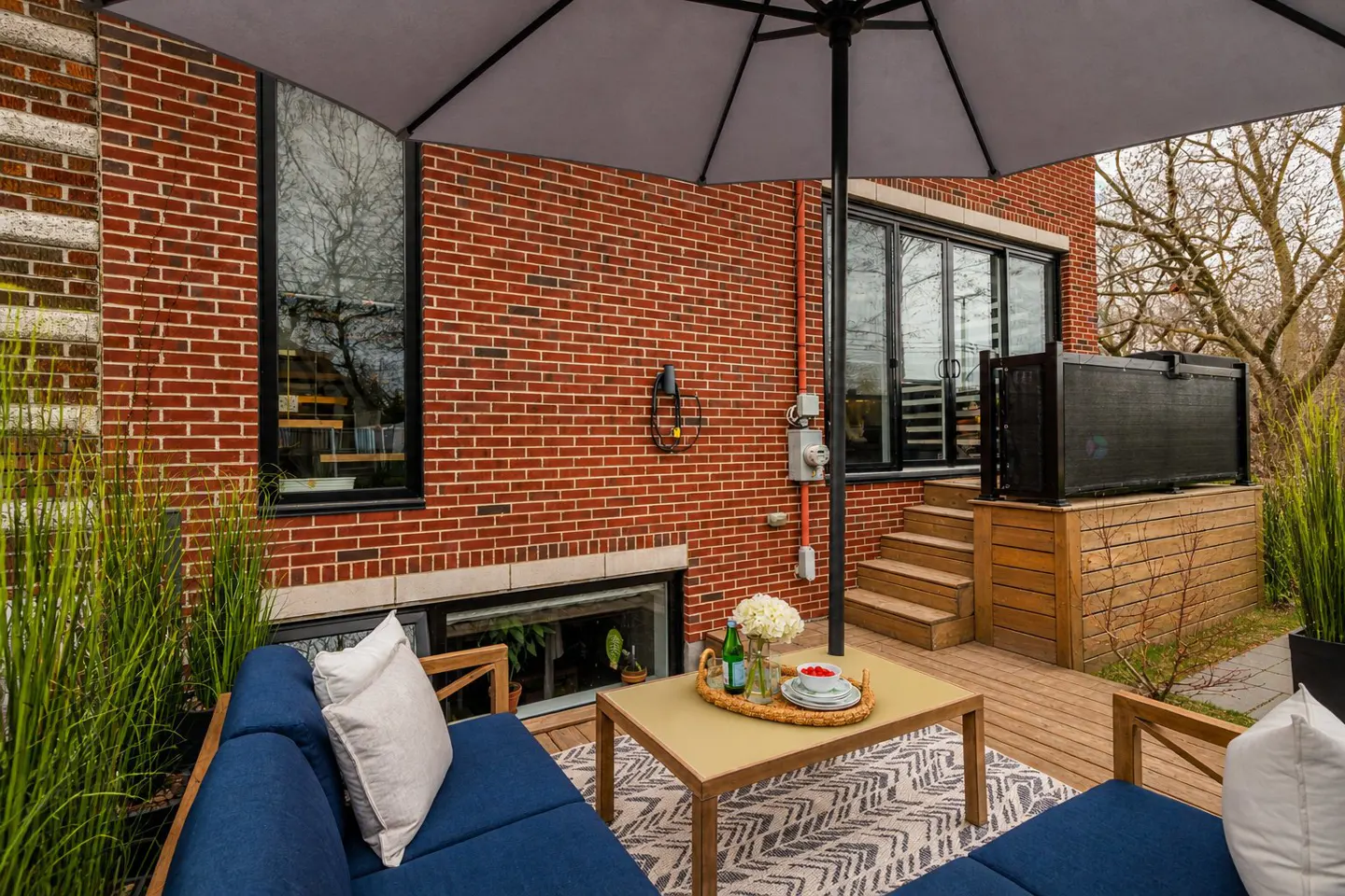 Outdoor patio with blue sofa, patterned rug, and table with flowers. Brick building with windows and stairs in the background.