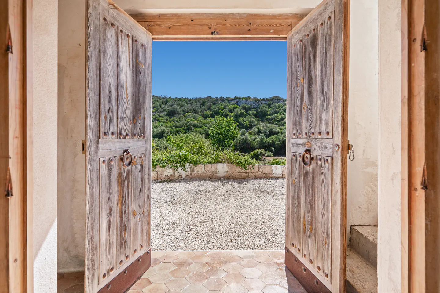 Open rustic wooden double doors reveal a gravel courtyard and a lush green hillside under a clear blue sky.