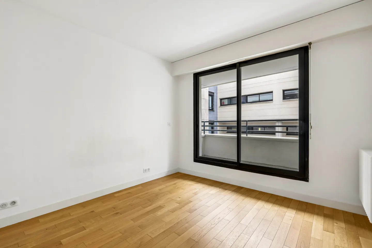 Bright, empty room with hardwood floors, white walls, and a large black-framed sliding glass door leading to a balcony.