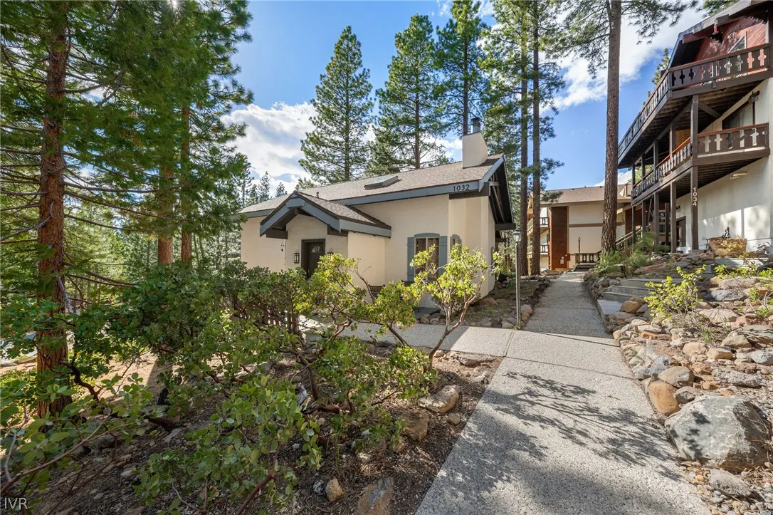 Exterior view of a beige house with a gray roof and trim, surrounded by tall trees and greenery. A stone pathway leads to the entrance.