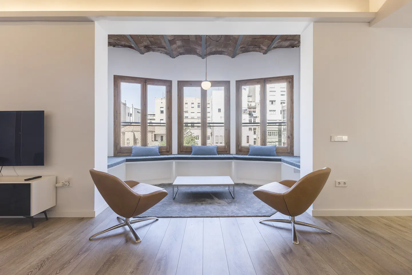 Living room with two tan chairs facing a window seat with blue cushions and a view of city buildings. Hardwood floors and a TV are visible.