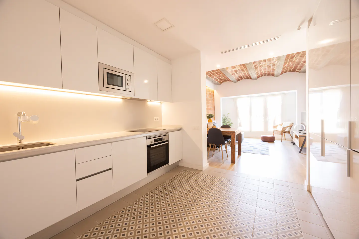 Bright, modern kitchen with white cabinets, stainless steel appliances, and patterned tile floor. Dining area and living room visible in the background.