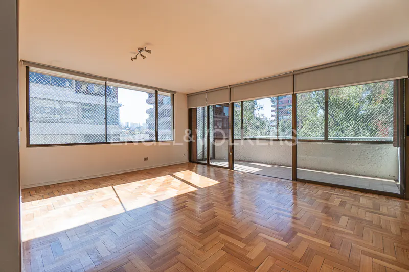 Bright, empty room with herringbone wood floors, large windows, and a balcony. Sunlight streams in, highlighting the Engel & Volkers logo.
