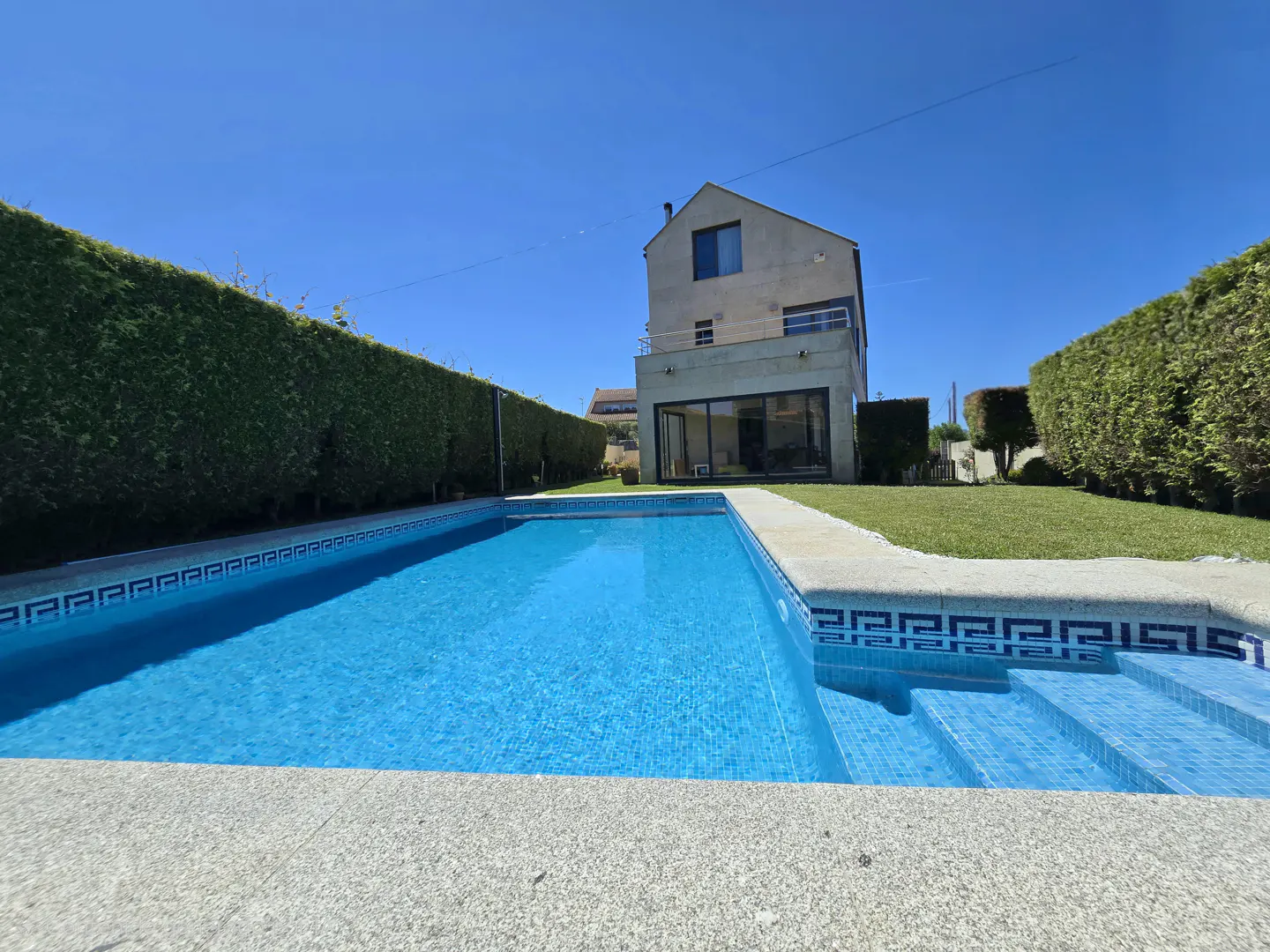 A bright blue swimming pool with tiled steps in a backyard with tall green hedges and a modern house under a clear sky.