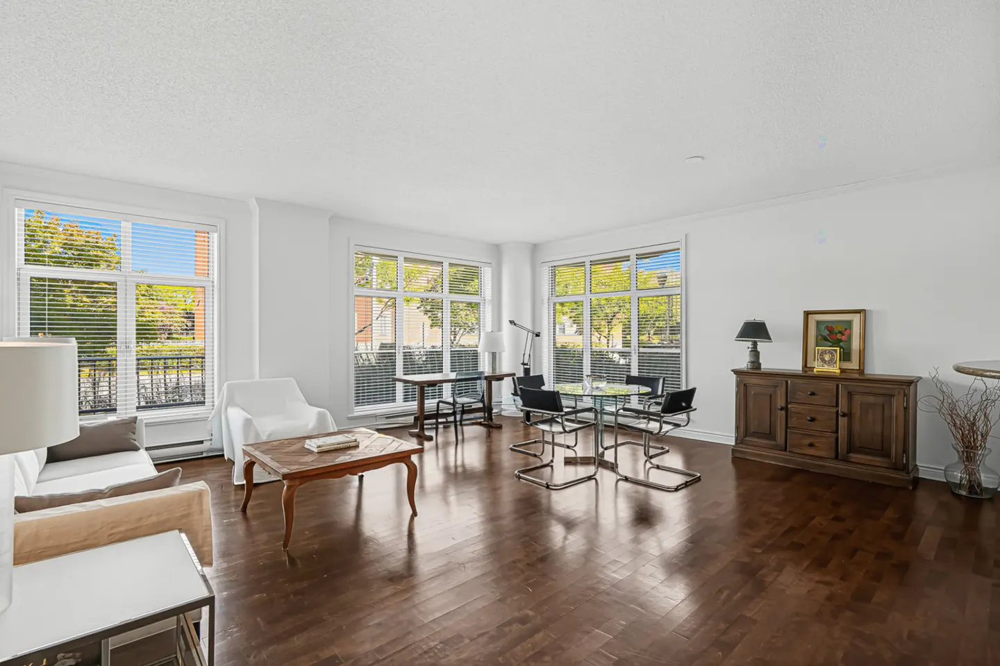 Bright living room with hardwood floors, white walls, and large windows. A sofa, coffee table, dining set, and wooden cabinet furnish the space.