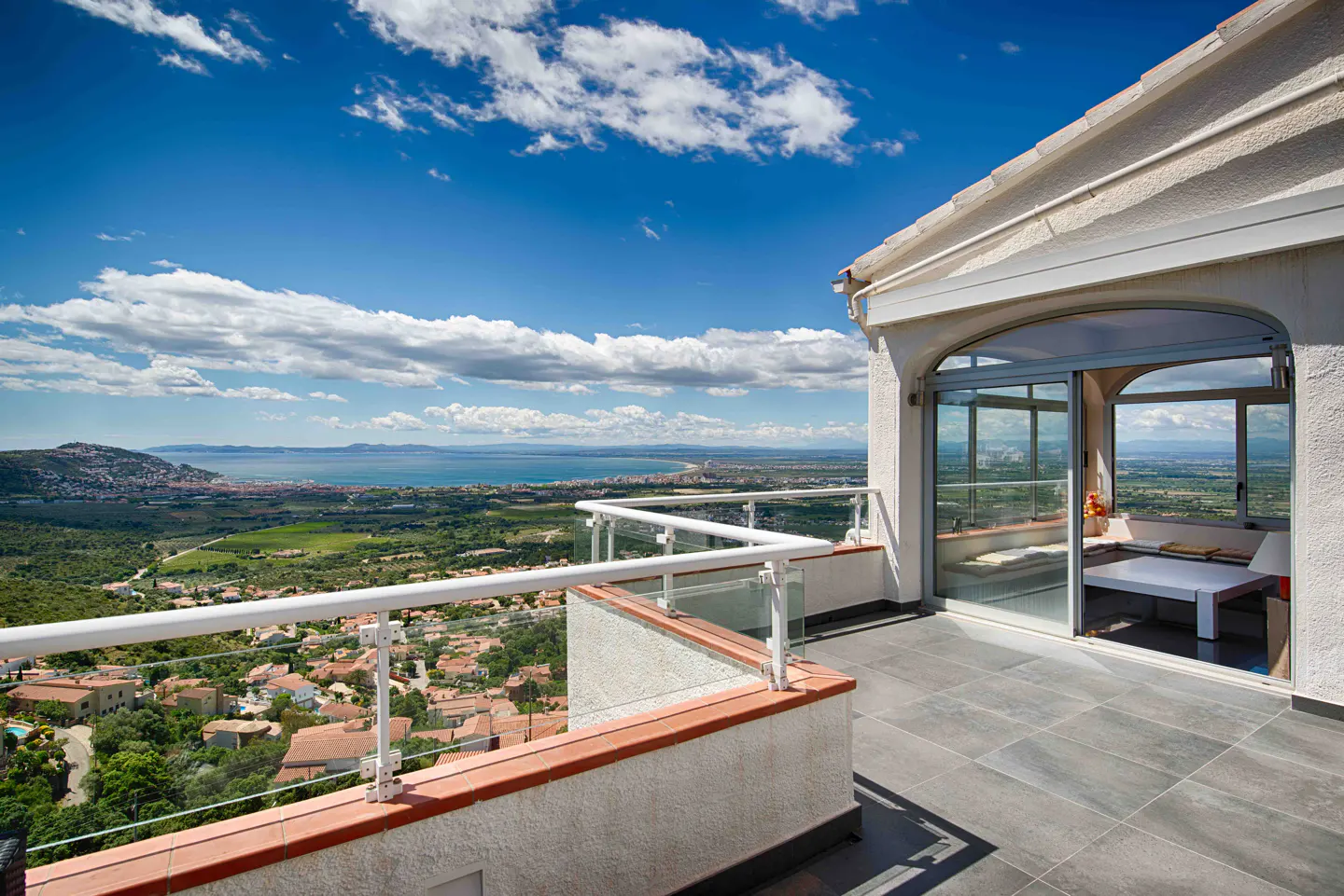 Balcony view of a white stucco house overlooking a town, green fields, and the ocean under a blue sky with white clouds.