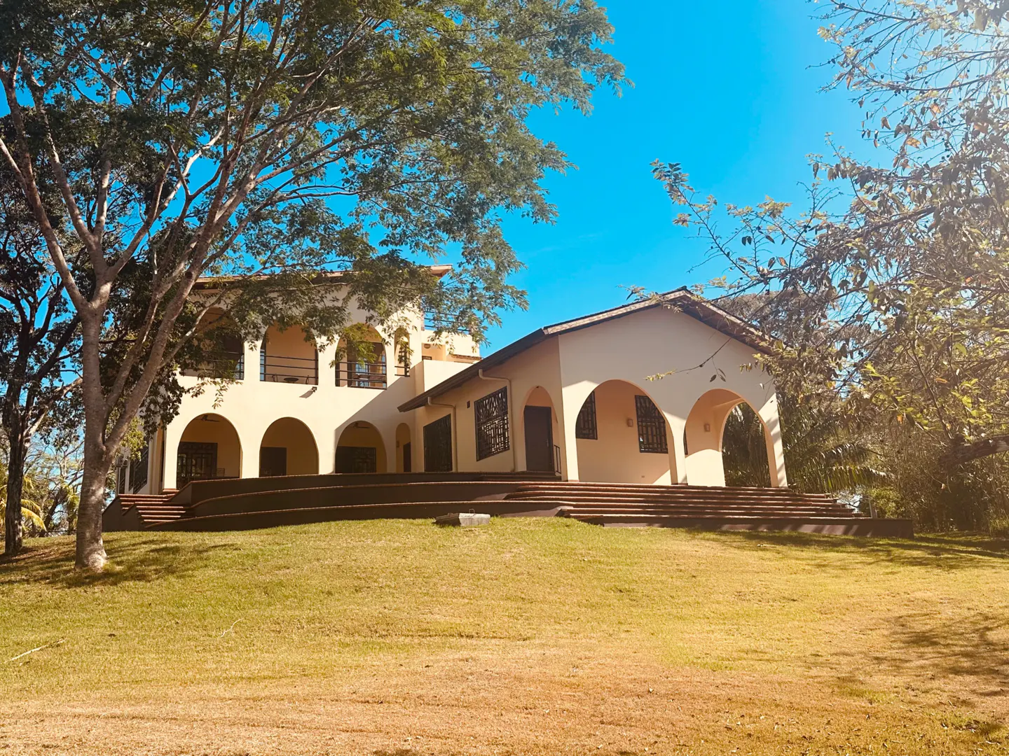 Two-story cream house with arched windows and balconies, set on a green lawn under a bright blue sky.