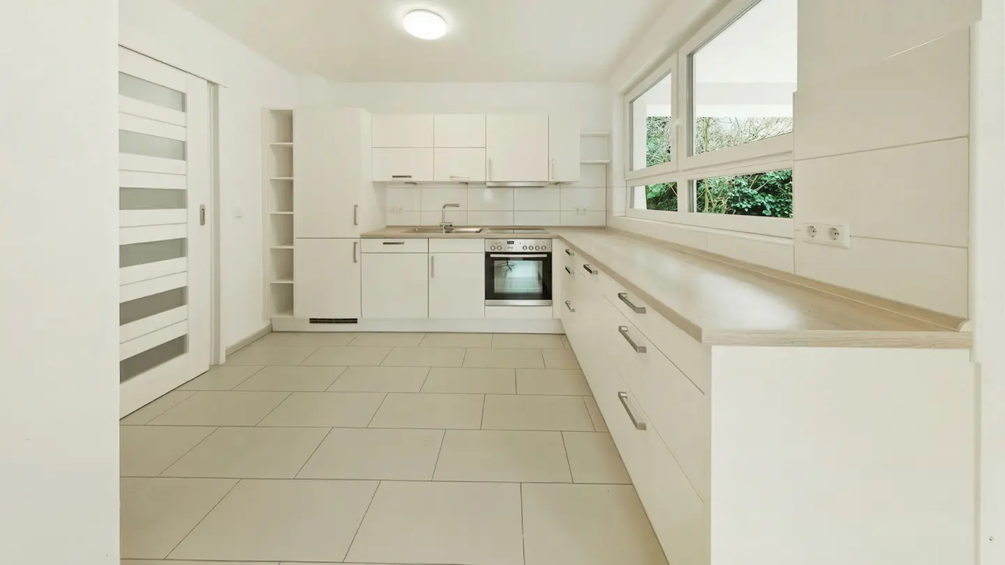 Bright, modern kitchen with white cabinets, light countertops, and tile flooring. A large window overlooks green foliage.