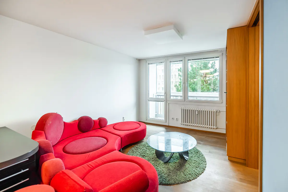 Bright living room with a red sectional sofa, a glass coffee table on a green rug, and a window view of trees.