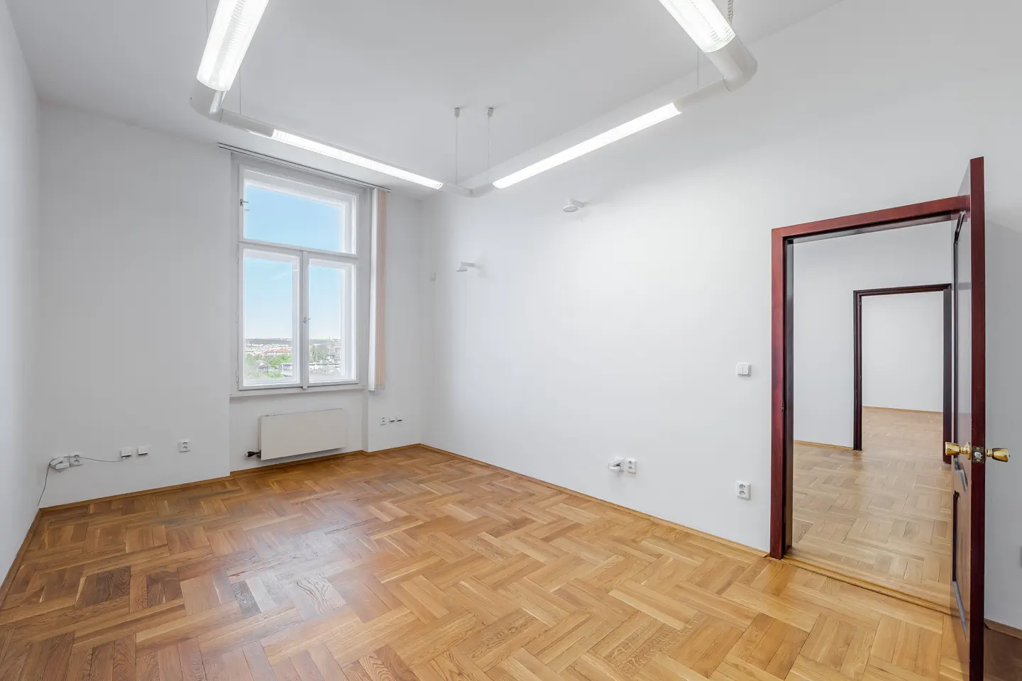 Empty room with herringbone wood floors, white walls, and a window with a city view. Ajar door shows another room.