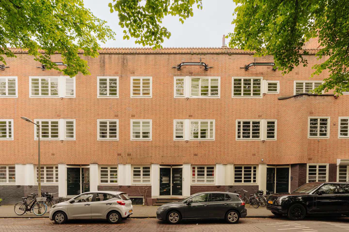 A three-story brick apartment building with white-framed windows and parked cars on the street.