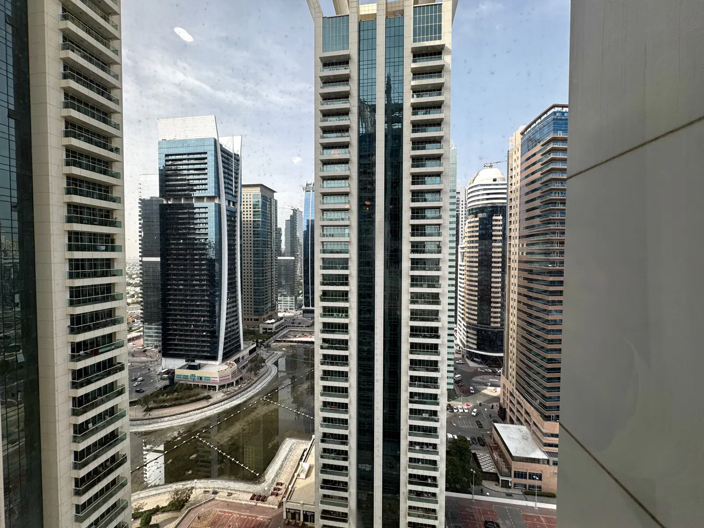 View from a high-rise window of a city skyline with modern buildings and a canal. Tall glass towers dominate the urban landscape.