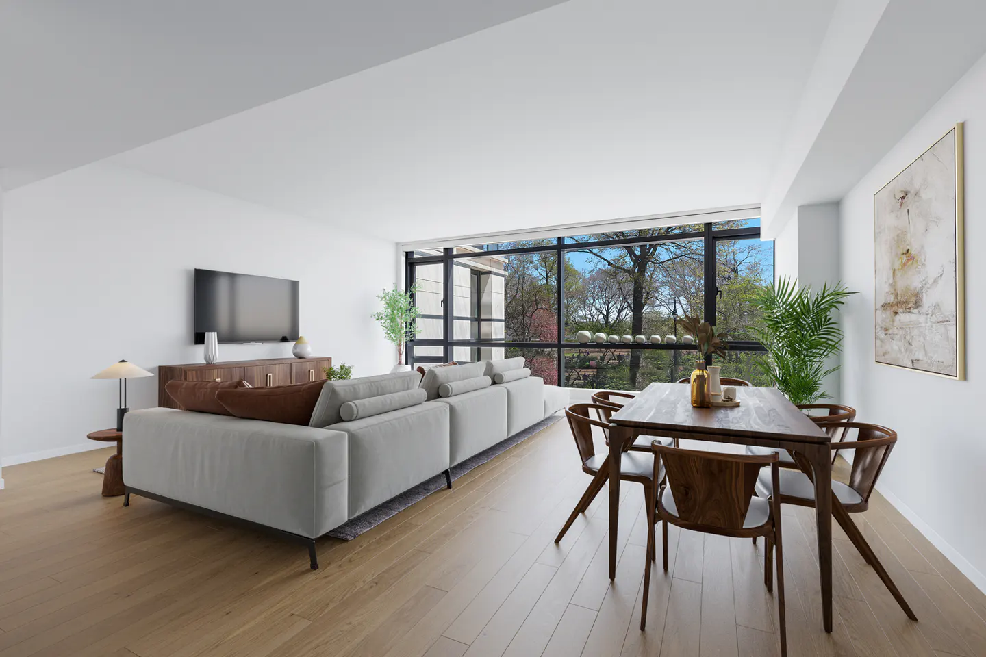 Bright, modern living room with a gray sofa, wood dining table, and large window overlooking trees. A TV hangs on the white wall.