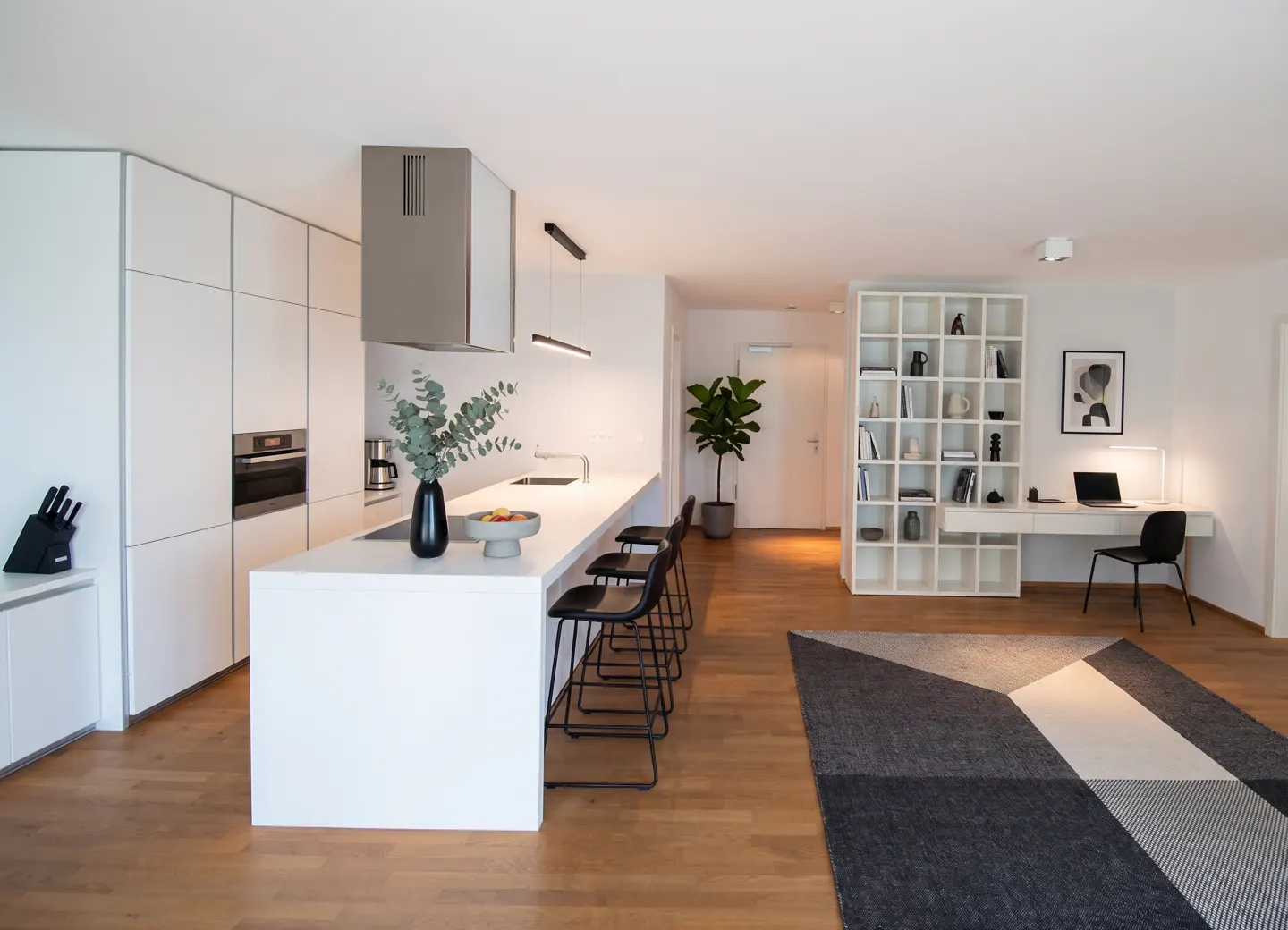 Bright, modern open-concept kitchen and living area with white cabinets, island with black stools, and a workspace with a white bookshelf.