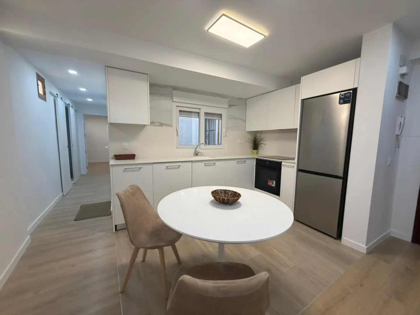 Bright, modern kitchen with white cabinets, stainless steel refrigerator, and round white table with two beige chairs. Hallway visible in background.