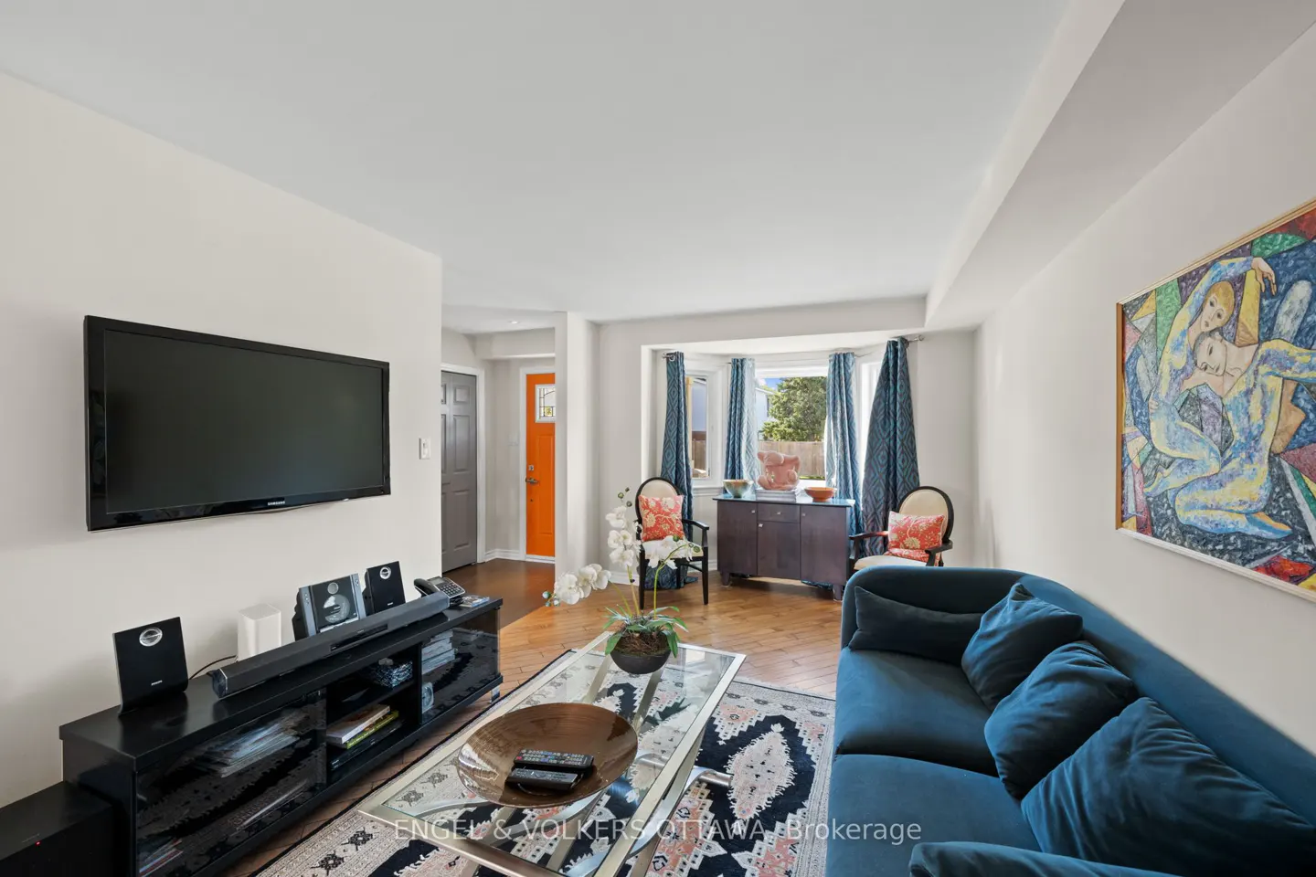 Living room with blue sofa, glass coffee table, and black media console under a wall-mounted TV. An orange door is visible in the background.