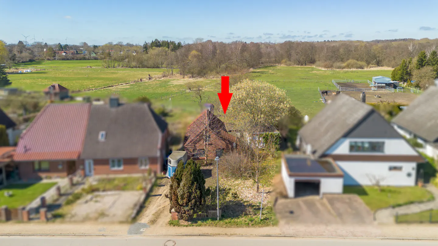 Aerial view of a house with a red arrow pointing to it, surrounded by trees and a green field.