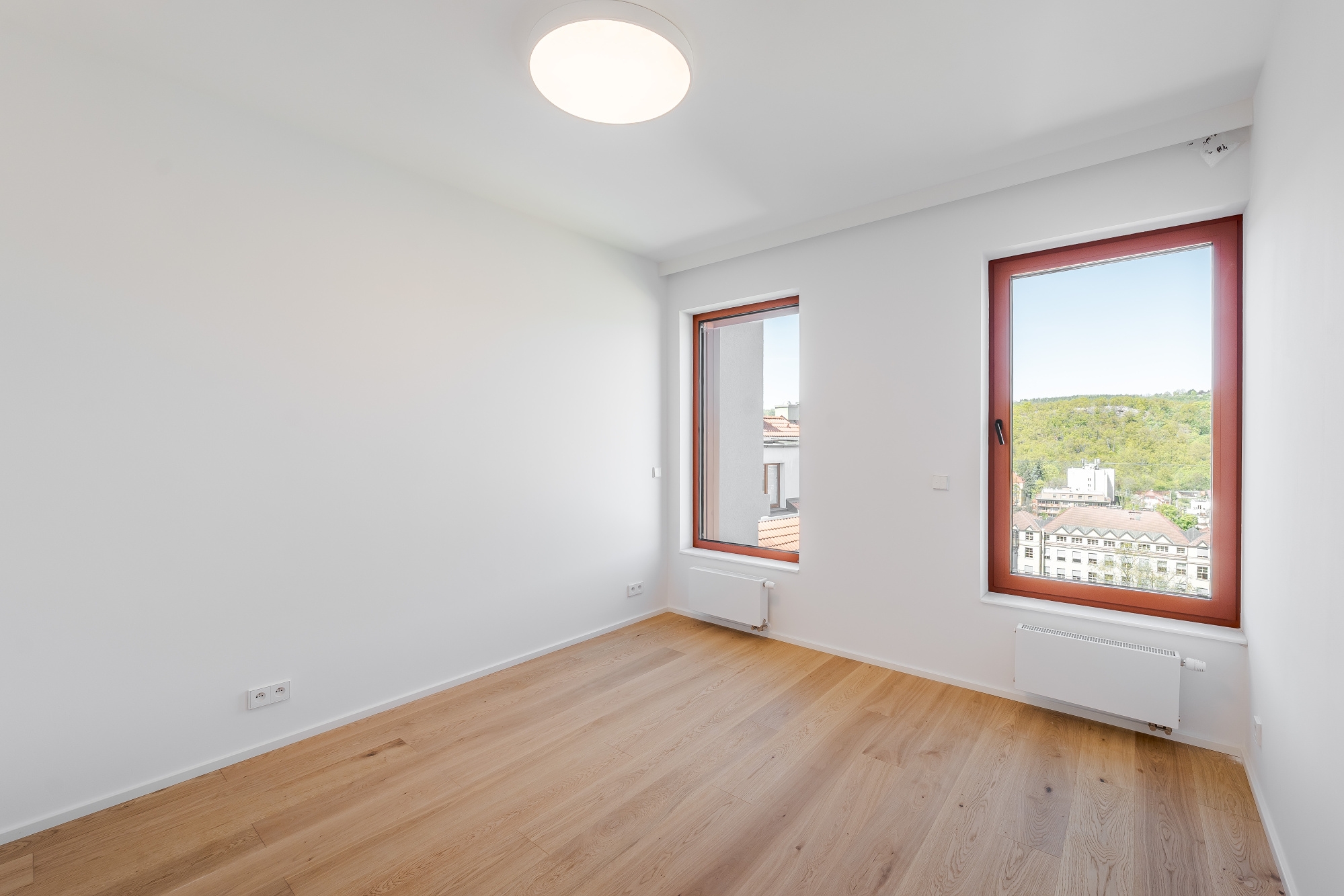 Empty room with white walls, light wood floors, a round ceiling light, and two windows with reddish-brown frames. [AI-generated text]