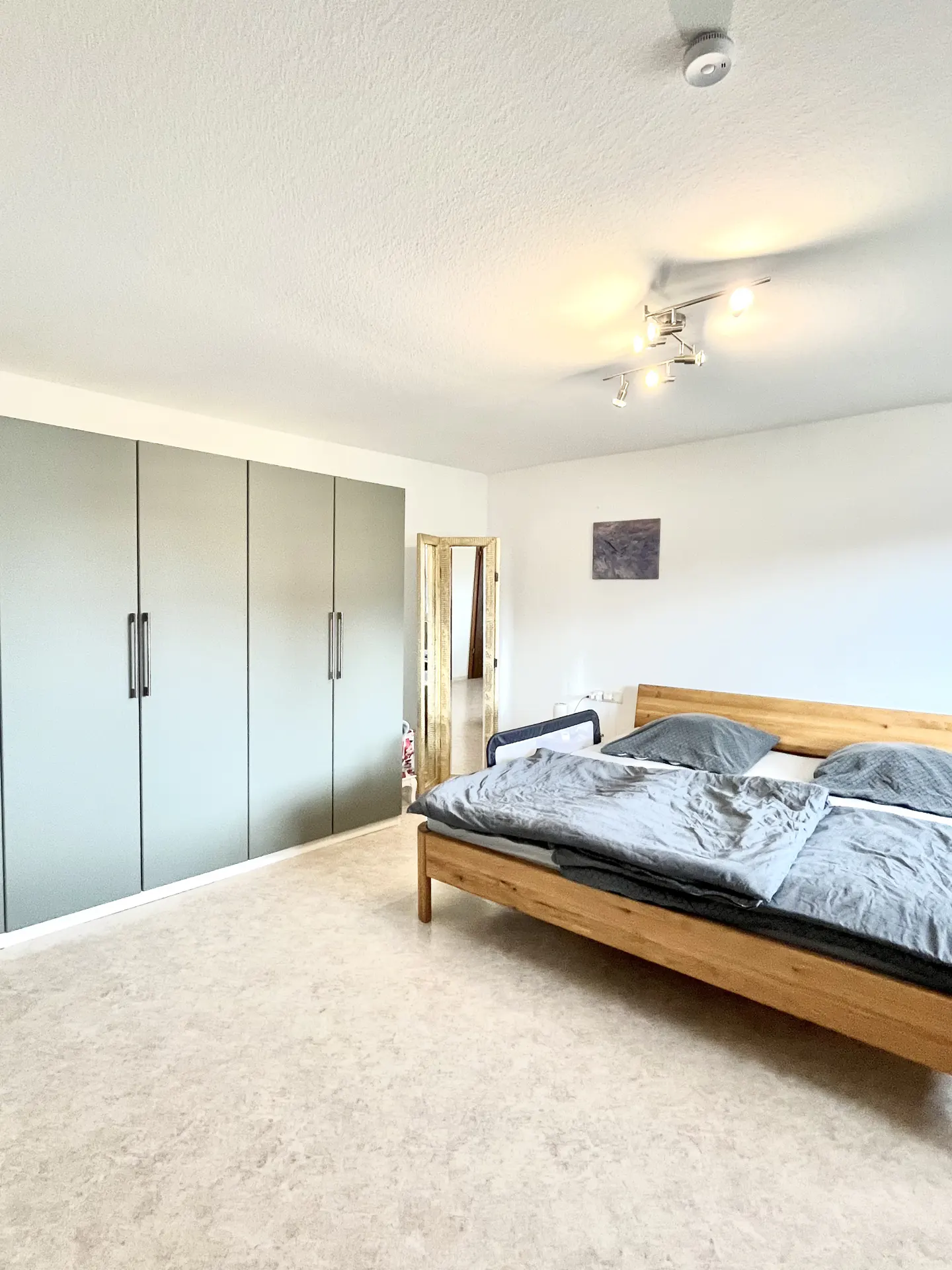 Bright bedroom with a wooden bed, gray bedding, and a wall of gray closets. A doorway leads to another room.