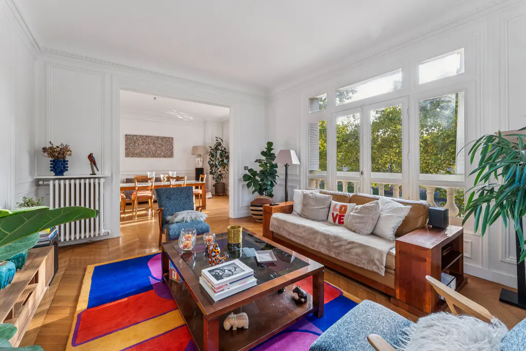 Bright living room with wood floors, white walls, and large windows. A colorful rug sits under a coffee table and sofa with "LOVE" pillow.