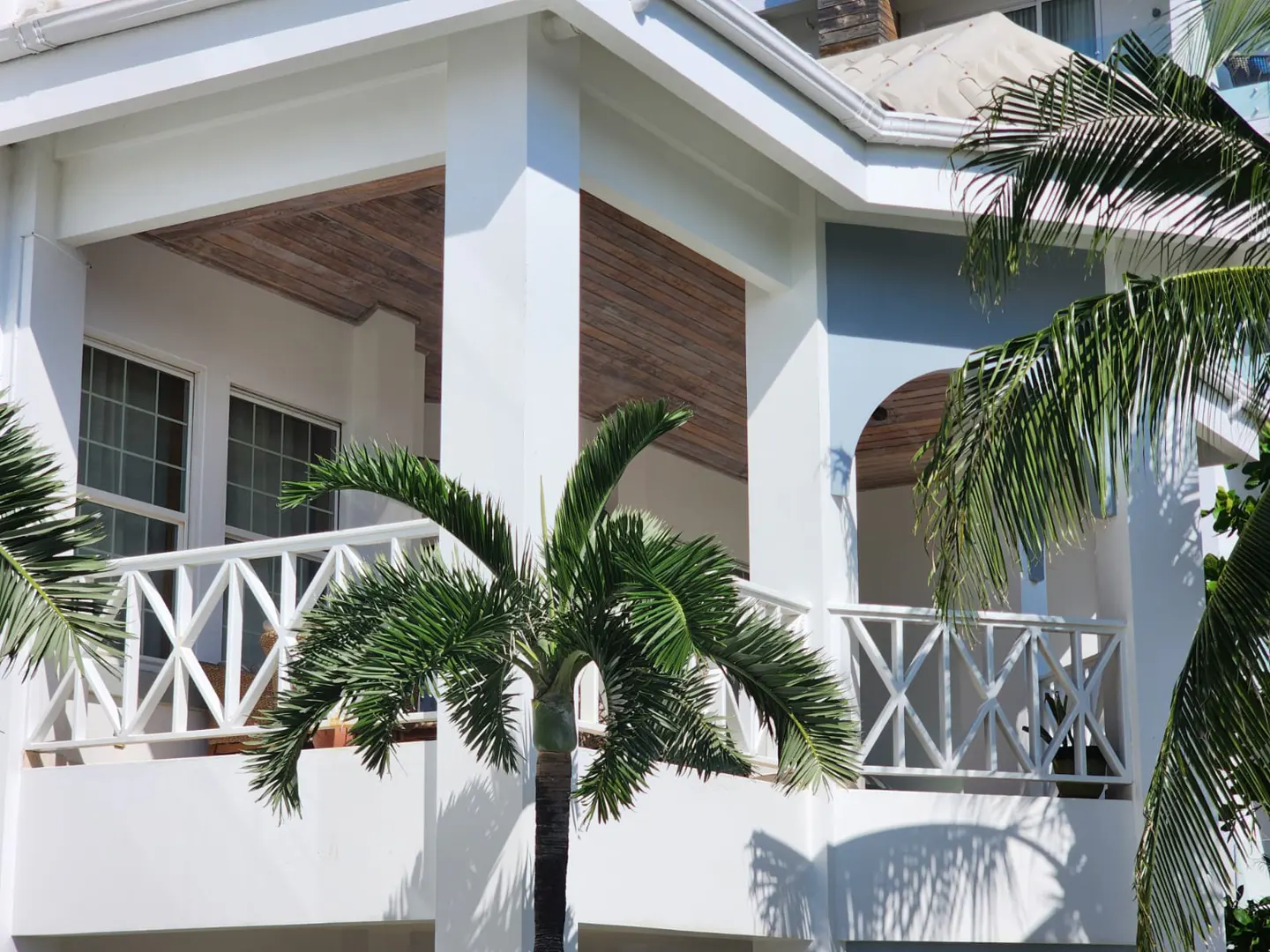 White building with balconies and palm trees. Balconies have white railings. The building has a light blue accent.
