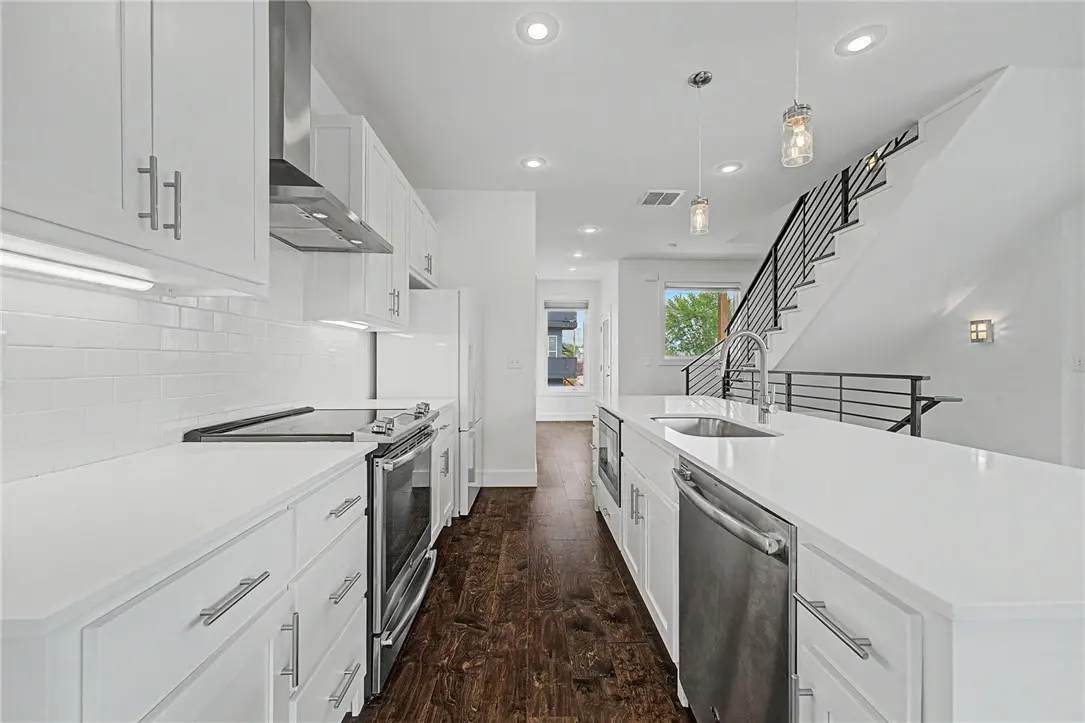 Bright, modern kitchen with white cabinets, stainless steel appliances, and dark wood floors. A staircase with black railings is visible.
