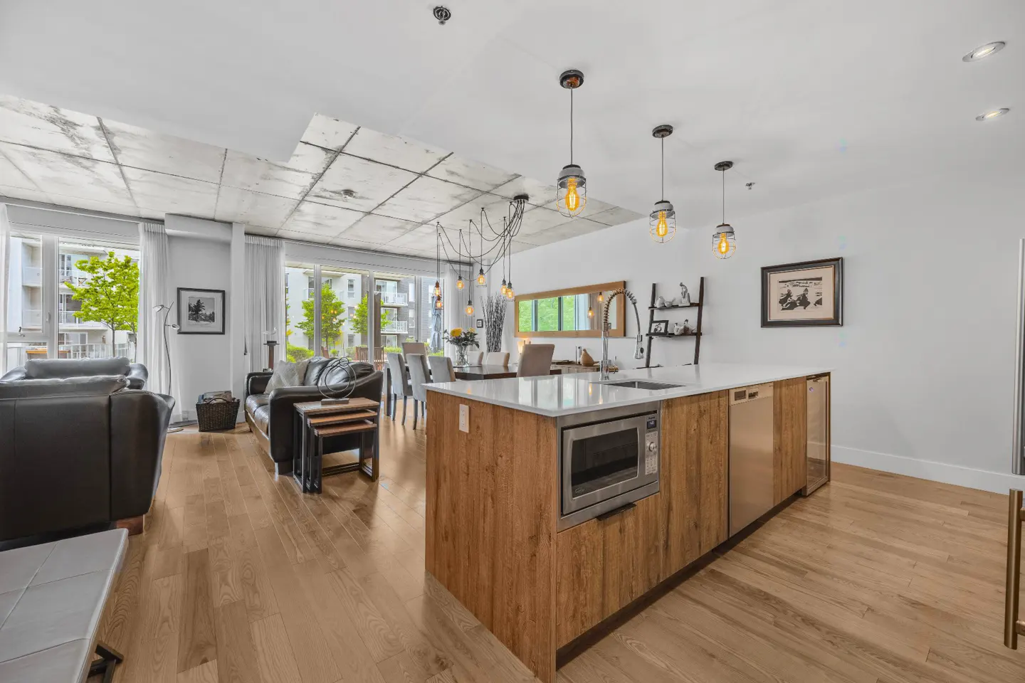 Open-concept living space with wood floors, leather sofas, and a kitchen island with stainless steel appliances. Pendant lights hang above.