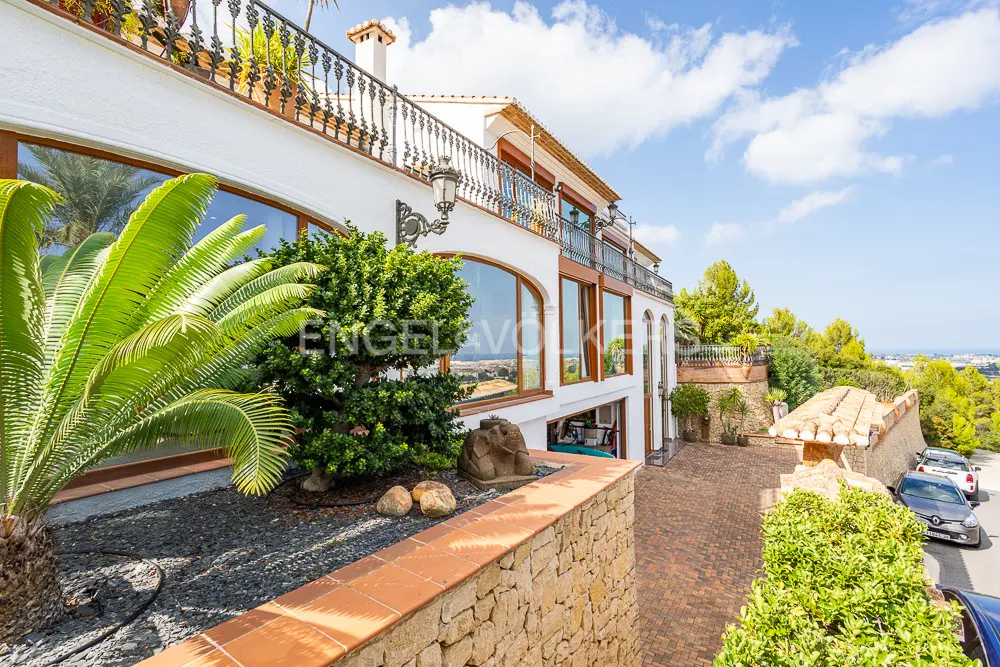 Exterior view of a white two-story house with large windows, a balcony, and a stone wall. Palm tree and greenery in the foreground.
