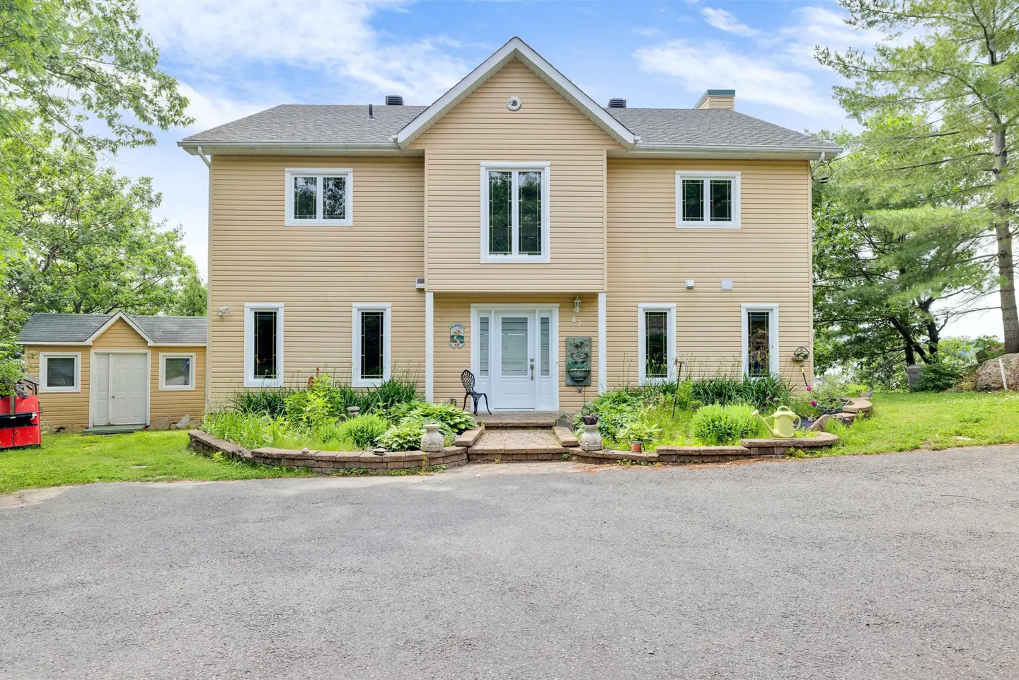 Two-story beige house with white trim, a white front door, and a gray roof. A small shed is on the left. The house is surrounded by greenery.
