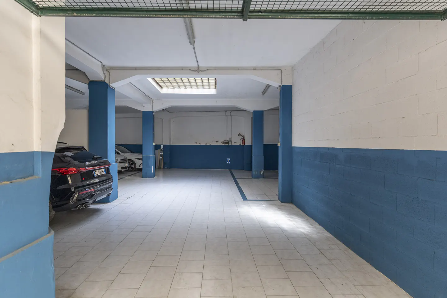 Indoor parking garage with blue pillars and a black car parked on the left. The walls are white and blue. Tiled floor.