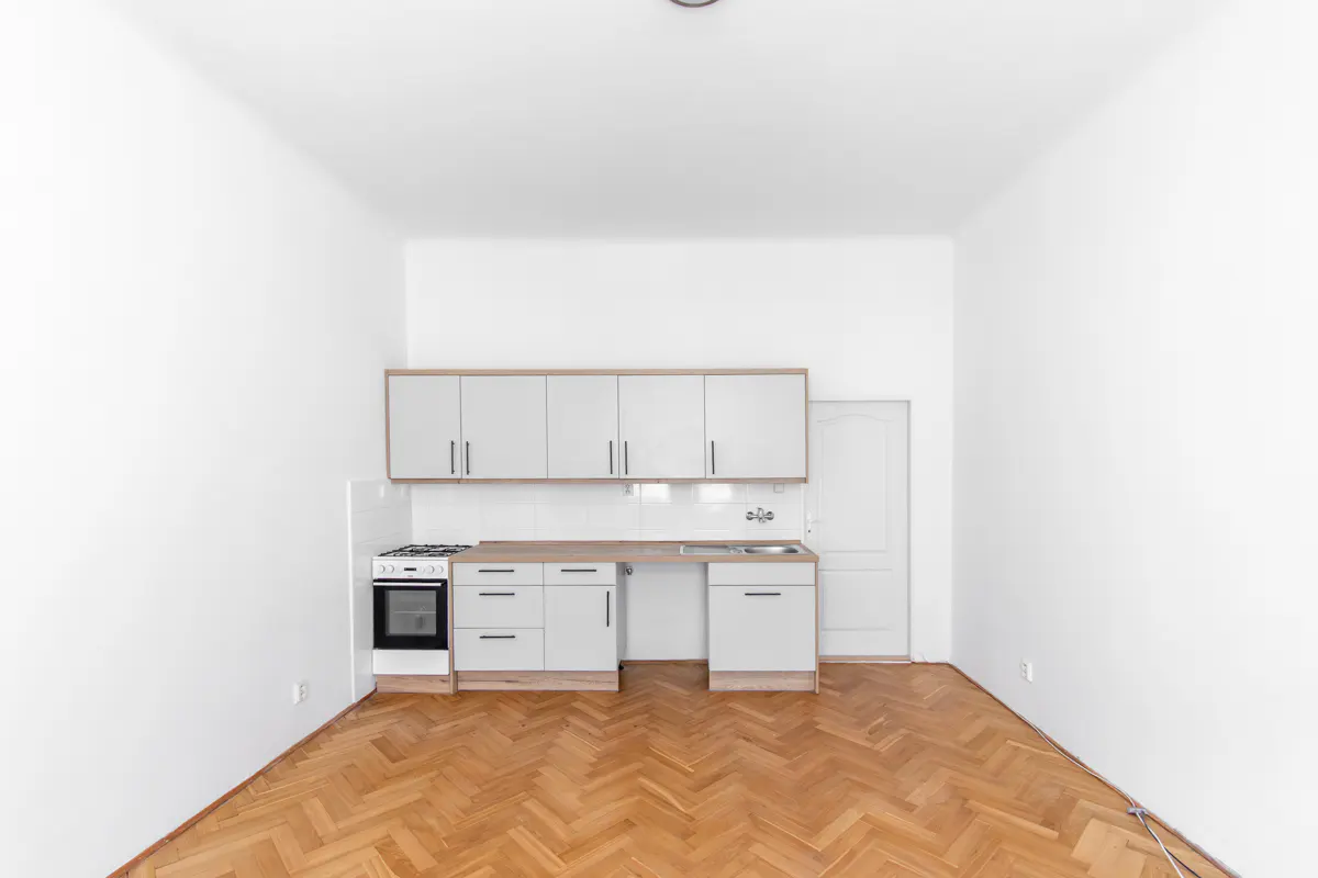 Bright, empty kitchen with white walls, herringbone wood floor, and white cabinets with a wood countertop. A white door is visible.