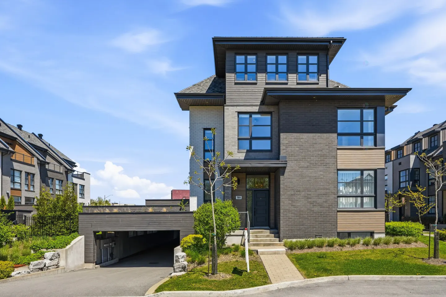 Modern two-story townhouse with gray brick, large windows, and a covered parking area on a sunny day.