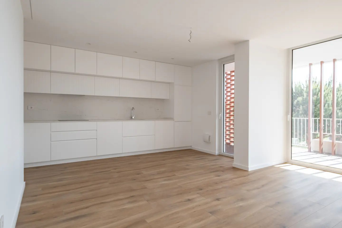 Bright, empty kitchen with light wood floors, white walls, and white cabinets. A sliding glass door leads to a balcony with trees visible.