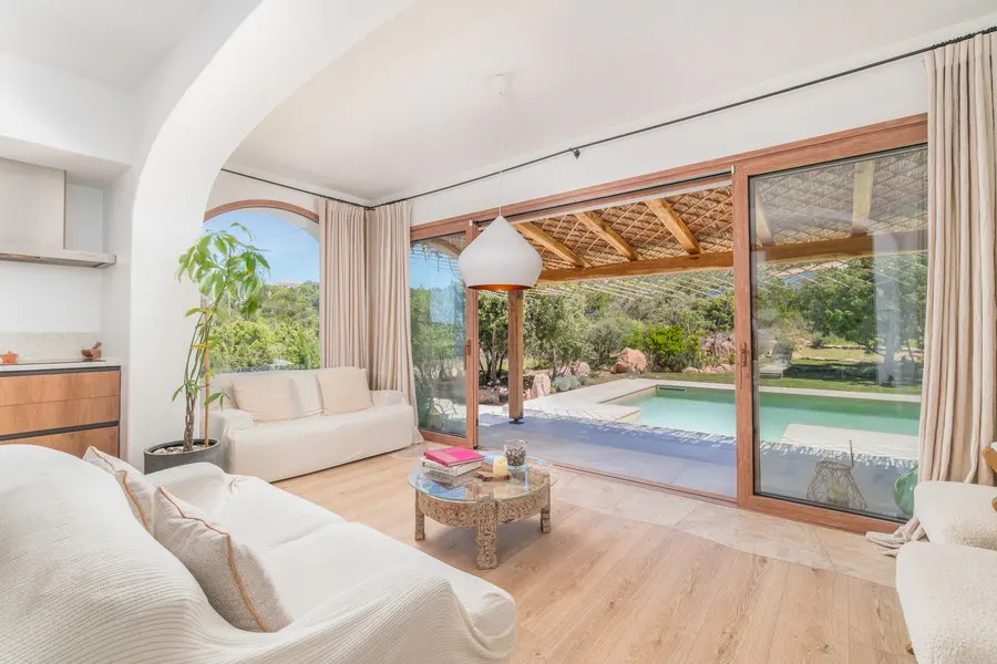 Bright living room with white sofas, wood floors, and a carved coffee table. Open sliding doors reveal a pool and lush greenery.