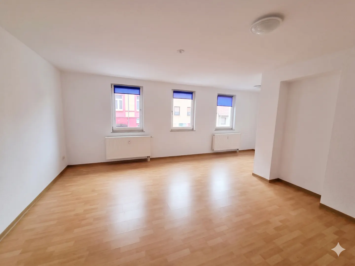 Empty room with white walls, light wood flooring, three windows with blue shades, and two white radiators.