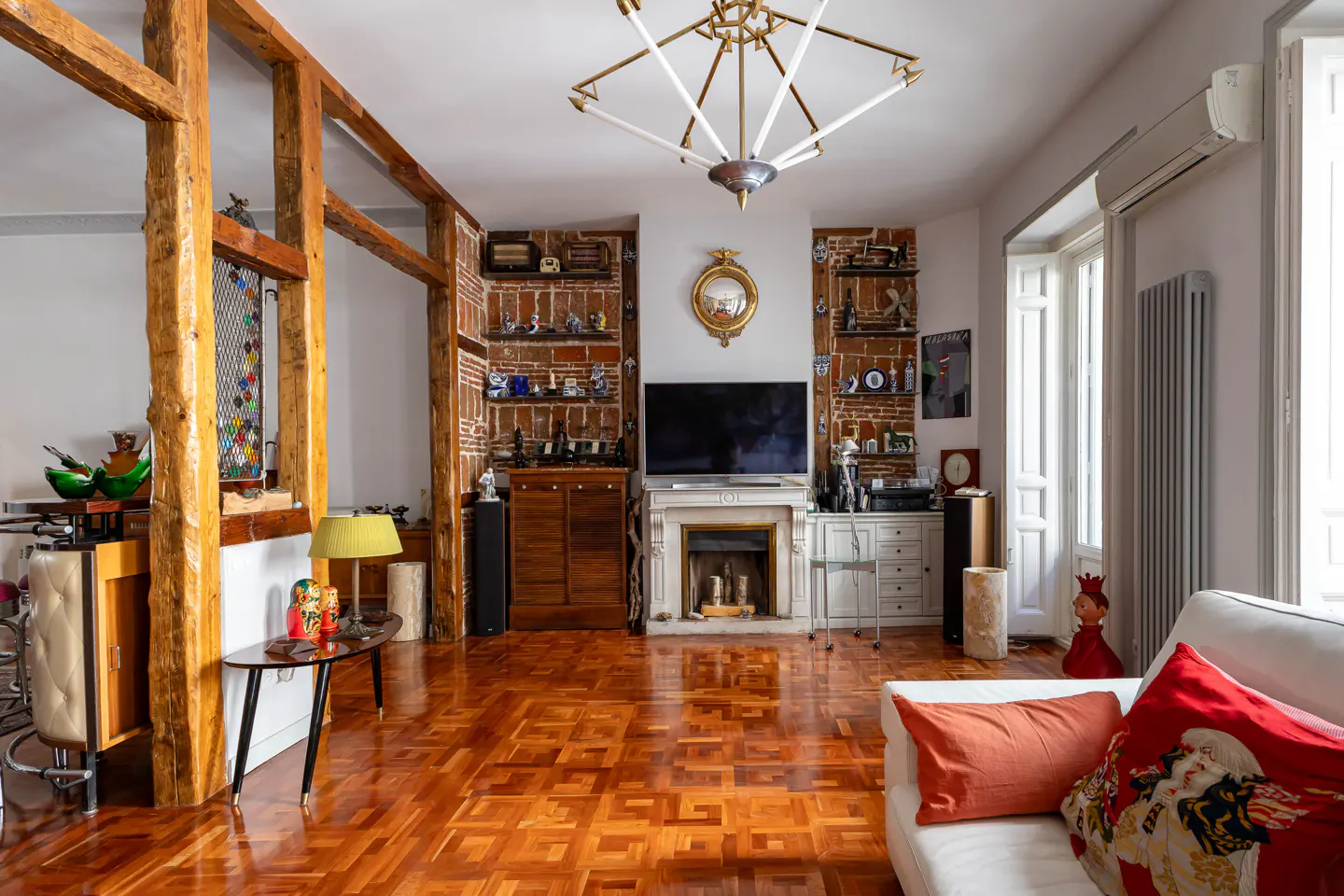 A living room with parquet floors, a fireplace, and exposed brick walls with shelves. A white sofa is in the foreground.