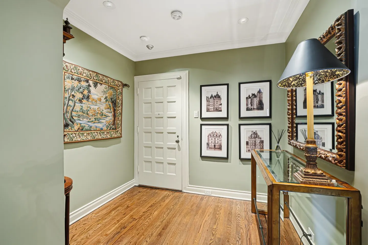 Entryway with light green walls, wood floors, and white door. A tapestry hangs on the left wall, and framed castle prints are on the right. A console table with a lamp sits against the right wall.
