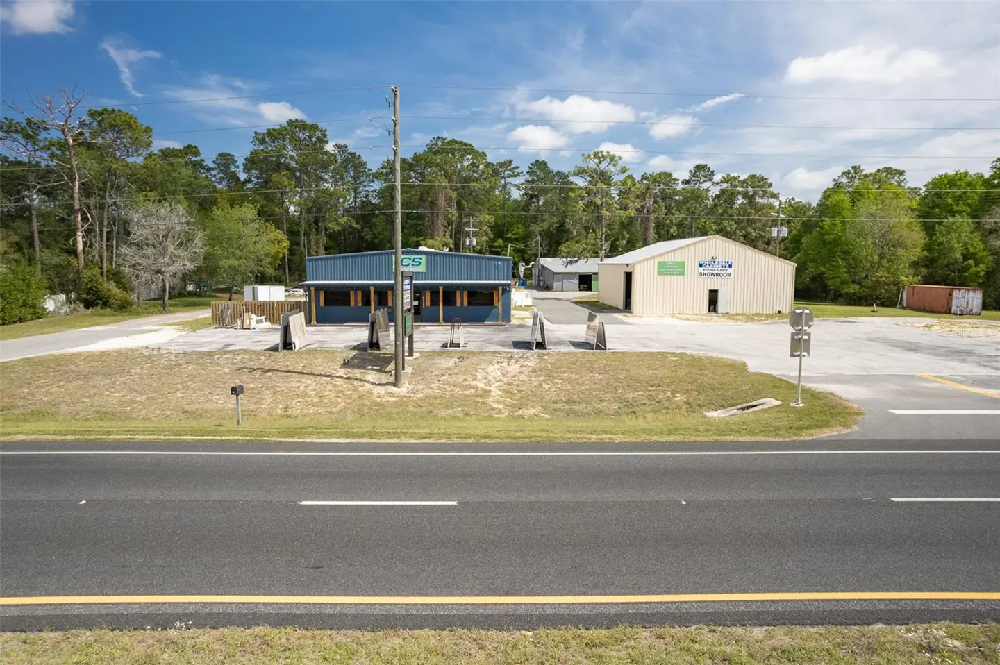 Commercial property with a blue building and a tan warehouse, set back from a black asphalt road with green trees in the background.