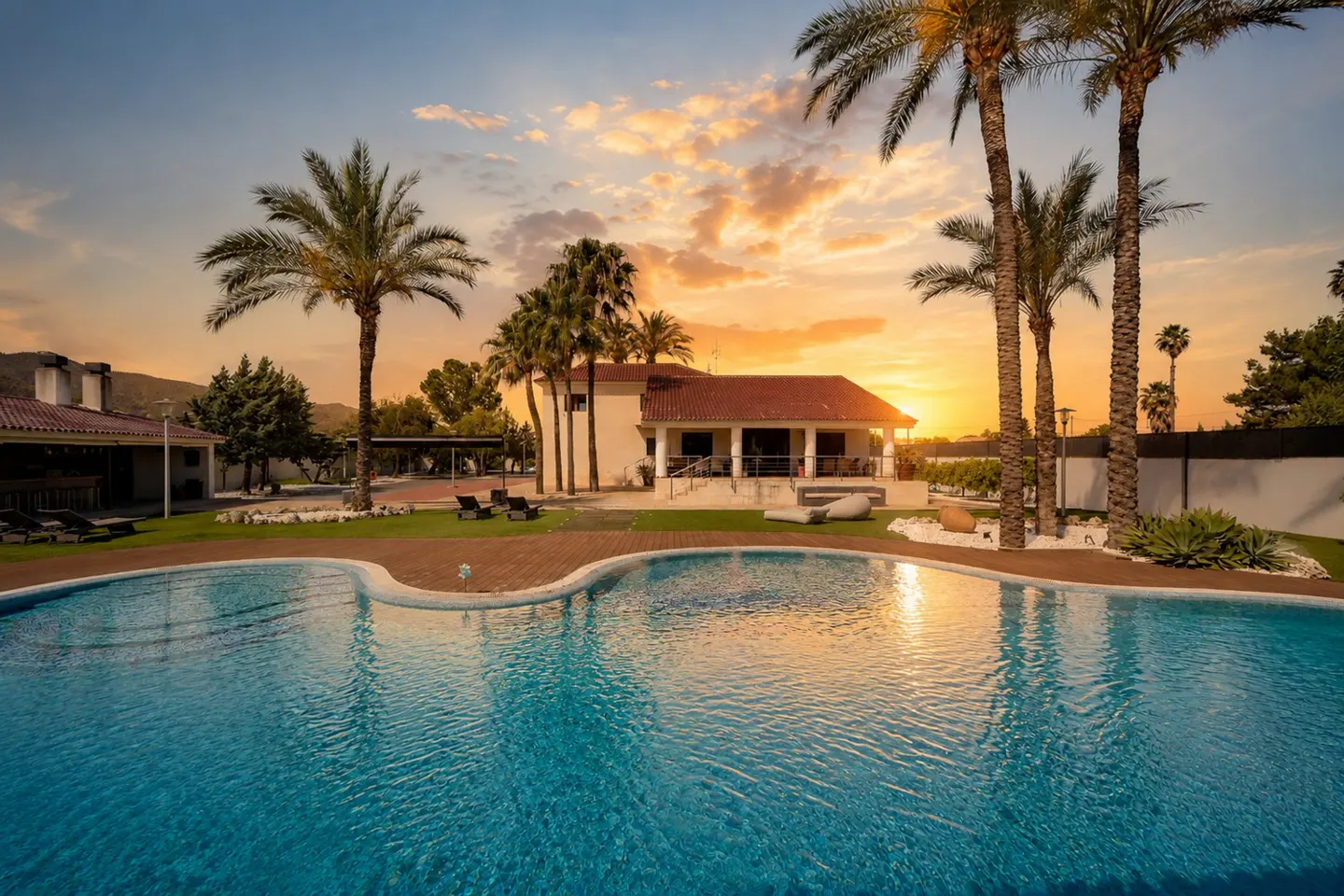 A luxurious home with a pool at sunset. Palm trees frame the white house with a red tile roof. The sky is orange and yellow.