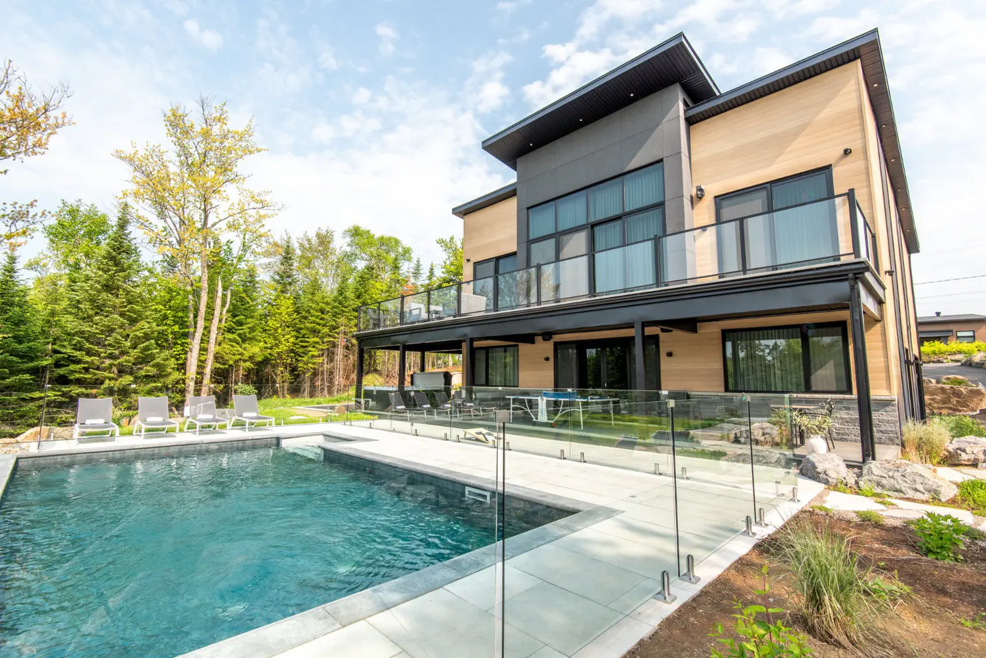Modern two-story house with a pool, deck, and glass railing. Lounge chairs line the pool. Trees and blue sky in the background.