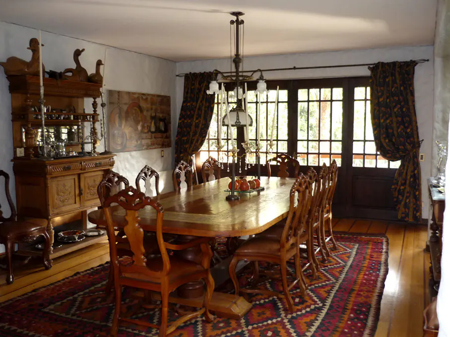 A dining room with a long wooden table, chairs, and a patterned rug. A hutch and a painting are on the left. Curtains frame a window.