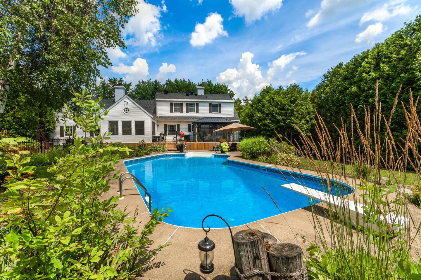 Backyard view of a white house with a blue in-ground pool, diving board, and lush green landscaping under a partly cloudy sky.