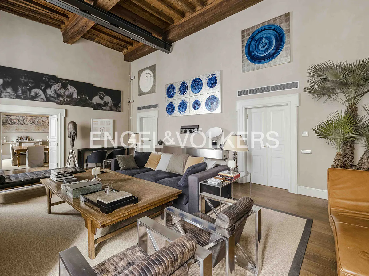 Living room with high ceilings, exposed wood beams, and modern art. Gray sofa, wood coffee table, and metal-framed chairs on a beige rug.